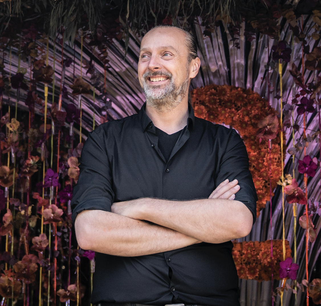 Tom De Houwer smiling with arms crossed in front of a floral installation